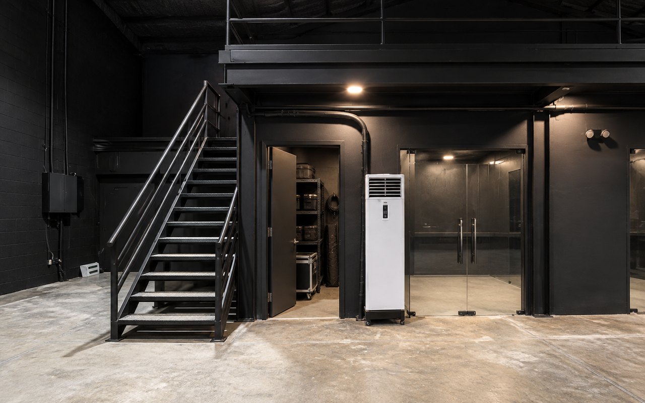 AI-staged visualization based on Studio W9 room photographs, showing the glass-fronted brand holding area beside the black steel staircase beneath the mezzanine.