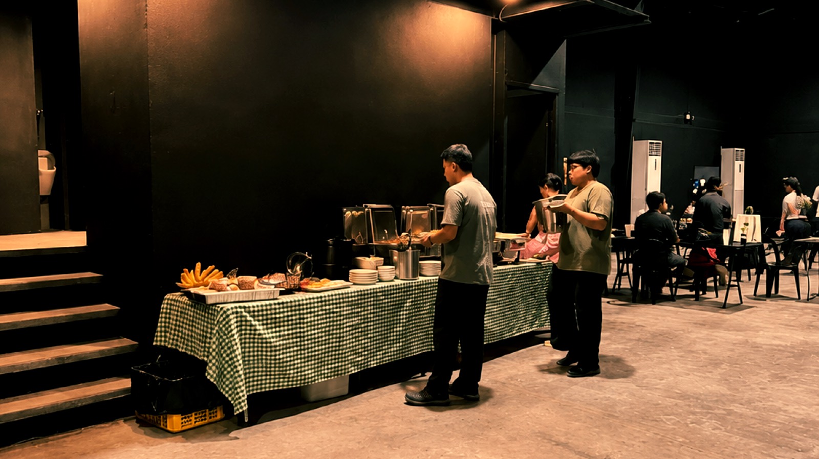 Catering buffet on Studio W9's open main floor — chafing dishes, breads, and plates on a green gingham-draped table with staff serving and guests dining at tables, against the blackout warehouse walls and polished concrete
